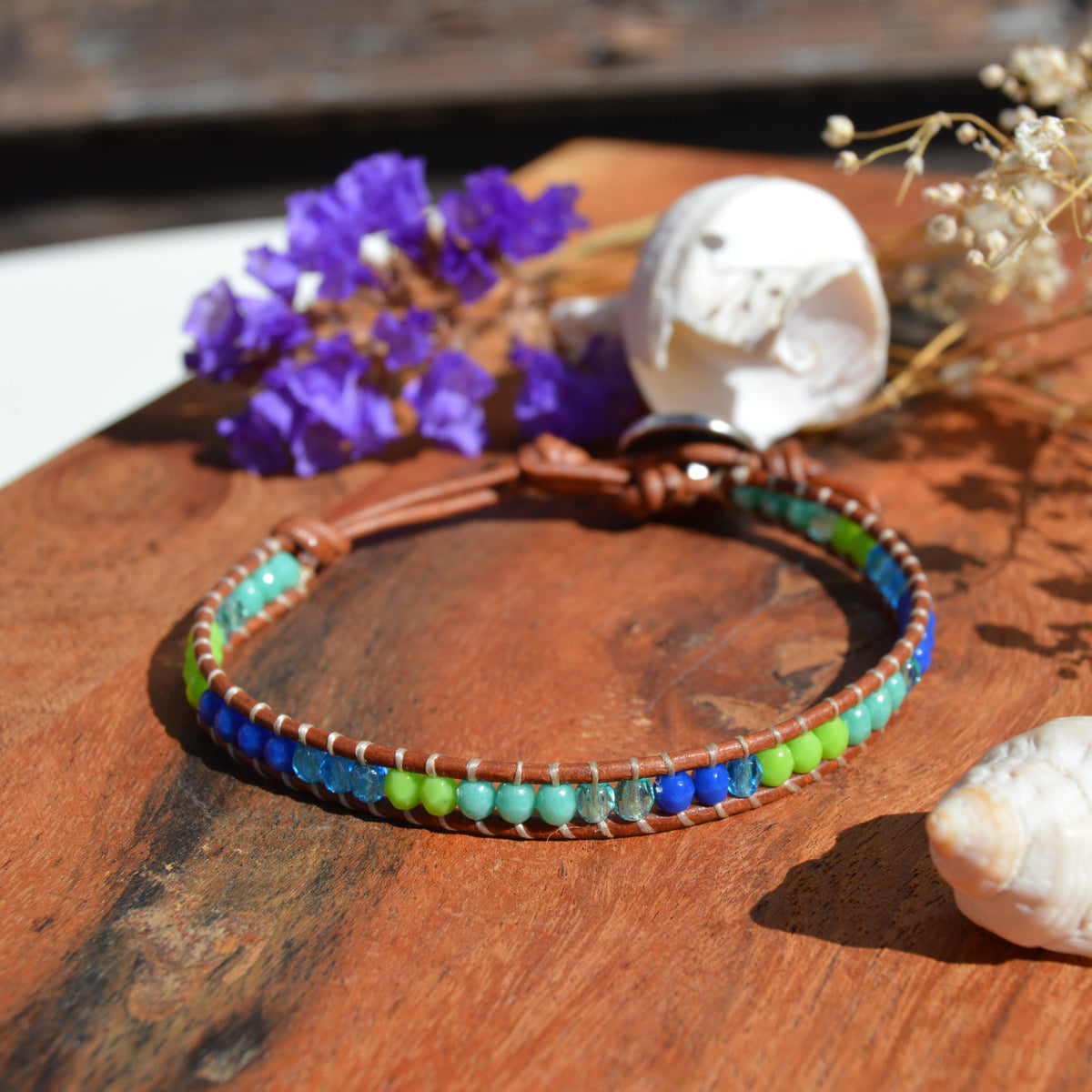 A handmade beaded bracelet with blue, green, and turquoise beads on a brown cord, metal button with tree of life design as a lock. Extension in leather.
