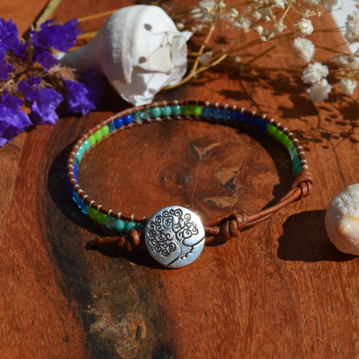 A handmade beaded bracelet with blue, green, and turquoise beads on a brown cord, metal button with tree of life design as a lock. Extension in leather.