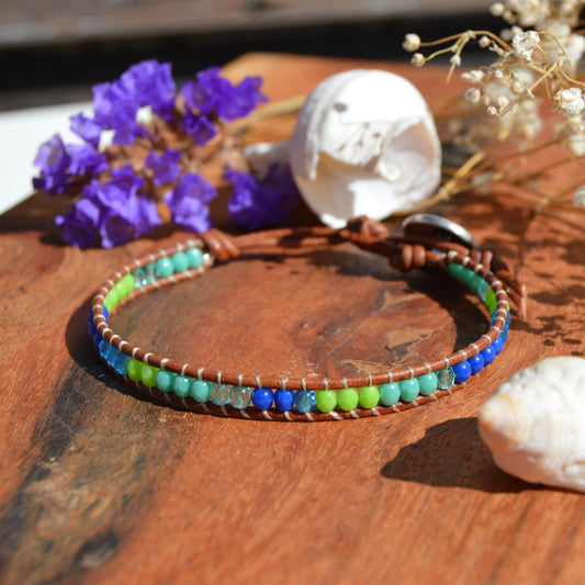 A handmade beaded bracelet with blue, green, and turquoise beads on a brown cord, metal button with tree of life design as a lock. Extension in leather.